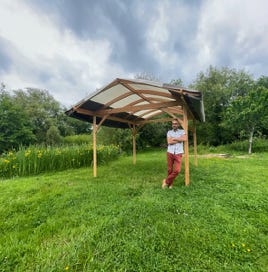 Tonnelle bois – vue de profil avec le fabricant accoudé à la structure. Artisanat local et convivialité sous un ciel nuageux.